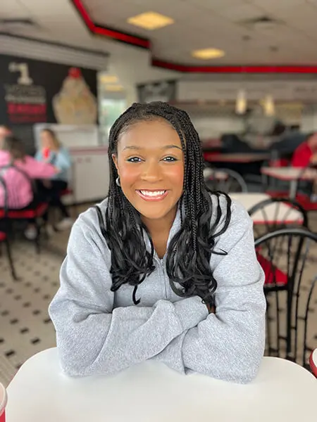 African American woman with long, thin braids and long-sleeve grey sweater poses for a headshot. Casual diner is in the background.