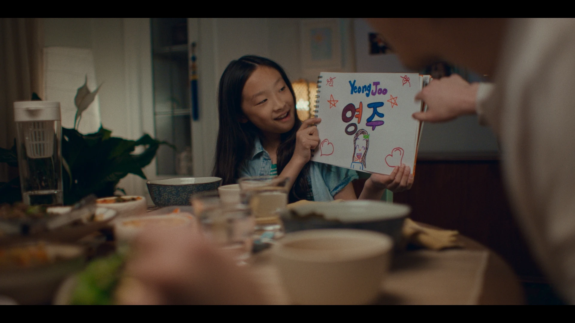 A yound Asian girl with long hair and a blue shirt sits at the dinner table with her family holding a drawing of her name “Yeong Joo.”
